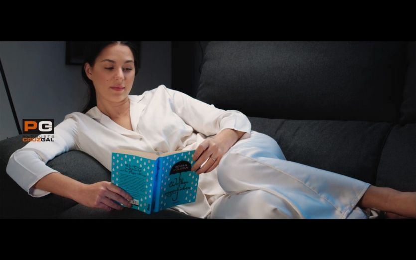 Fotograma de anuncio de televisión para la firma CruzGal. Vemos a una chica en pijama leyendo relajadamente en el sofá de su salón