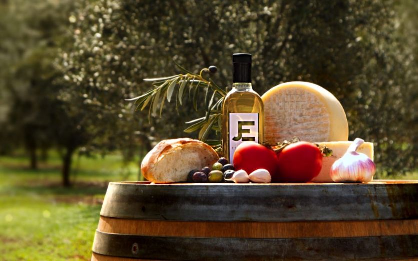 Fotografía. Bodegón de diferentes productos de la Finca Escudero y de los productores vecinos: pan, tomates, ajos, aceitunas, queso y aceite. Tomada en la misma finca con los olivares al fondo.